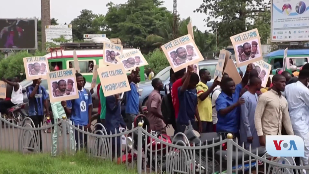 Eine Gruppe von Menschen hält Schilder in Händen und steht vor einem Zaun, umgeben von Fahrzeugen, Bäumen, Masten, Plakatwänden und einem Bildschirm. Im Hintergrund ist Gras und der Himmel zu sehen, und es scheint eine Regierungentscheidung gegen Video-Konferenzen zu protestieren.