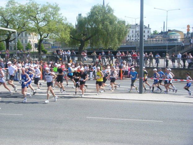 Gruppe von Läufern bei einem Marathon auf einer Straße mit Zielpfosten, Band und Zuschauern, umgeben von Absperrungen und Pfosten unter einem bewölkten Himmel.