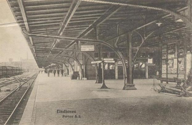 Ein Schwarz-Weiß-Foto eines Bahnhofs mit Menschen auf dem Bahnsteig, Eisenbahnschienen links, einem Zug auf der rechten Seite und Gebäuden im Hintergrund, mit dem Text "Eindhoven Post Office, Berlin, Germany" unten.