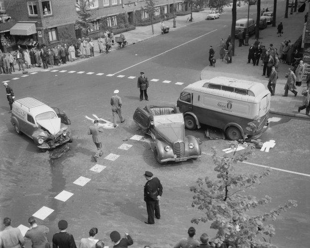 Eine Schwarz-Weiß-Szene eines Autounfalls an einer Straßenkreuzung, mit mehreren Fahrzeugen einschließlich eines Lieferwagens, einer Gruppe von Menschen, die sich darum versammeln, und Gebäuden, Bäumen, Pfählen und Texttafeln im Hintergrund.