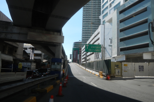 Stadtstraße mit Verkehrskegeln, Fahrzeugen, einer Brücke auf der linken Seite, Gebäuden mit Schildern, Laternenpfählen und einem Himmel im Hintergrund.