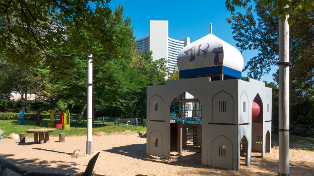 Ein Spielplatz mit einem zentralen Spielgerüst, umgeben von Bäumen, Pfählen, Bänken und anderen Gegenständen, mit Gebäuden und einem klaren blauen Himmel im Hintergrund.