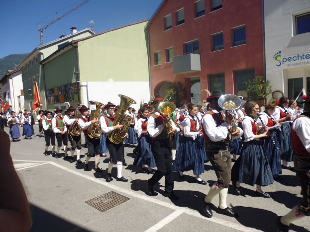 Menschen in traditioneller bayrischer Tracht spielen Instrumente, während sie eine Straße entlanggehen, die von Gebäuden gesäumt ist, einige halten Fahnen, mit einem Hügel und einem klaren Himmel im Hintergrund.