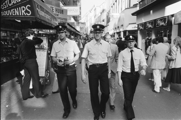 Drei Polizisten in Uniform gehen eine belebte städtische Straße mit hohen Gebäuden und Geschäften entlang, mit Passanten und einem sichtbaren Himmel im Hintergrund.