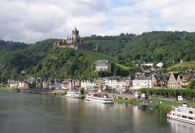 Ein malerischer Blick auf den Rhein in Deutschland mit einer Burg auf einem Hügel, Booten auf dem Fluss, Fahrzeugen auf der Straße und einem bewölktem Himmel.