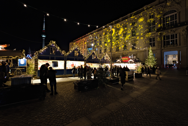 Ein lebendiger Weihnachtsmarkt in Berlin, Deutschland, mit Menschen um bunt beleuchtete Stände versammelt, festliche Dekorationen und Gebäude mit Fenstern im Hintergrund unter einem dunklen Himmel.