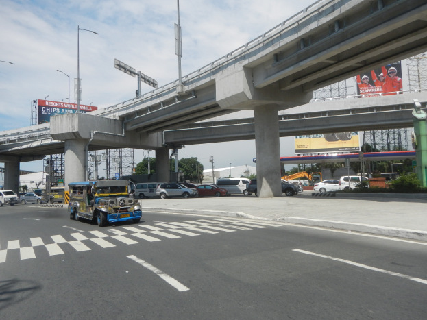Vielbefahrene Hauptstraße mit Fahrzeugen, Zebrastreifen, Überführung mit Pfeilern, Laternen, Informationsschilder, Bäume, Gebäude und bewölkter Himmel.