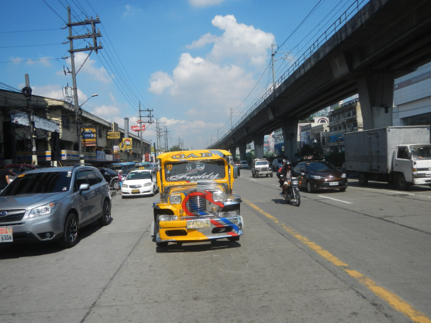Ein gelbes Taxi fährt auf einer Straße mit hohen Gebäuden, Strommasten mit Drähten und einer Brücke im Hintergrund bei bewölktem Himmel, mit einem Wasserzeichen.