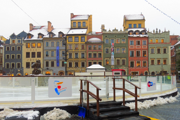 Eisbahn in einem Stadtplatz umgeben von Gebäuden, Treppen, Geländern, Informationsschildern, Laternen, Bäumen und schneebedecktem Boden unter einem Himmel.