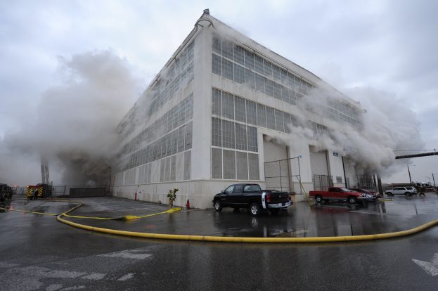 Ein großes Industriegebäude steht in Flammen mit dickem Rauch, umgeben von Fahrzeugen und ein paar Schaulustigen, vor einem bewölkten Himmel.