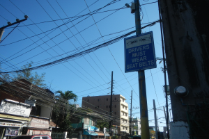 Stadtstraße mit fahrenden Autos, Strommäste mit Drähten, Gebäude mit Fenstern, Bäume, Namensschilder und ein "Fahrer Müssen Sitzgürtel Tragen"-Schild an einem Strommast vor einem sichtbaren Himmel.