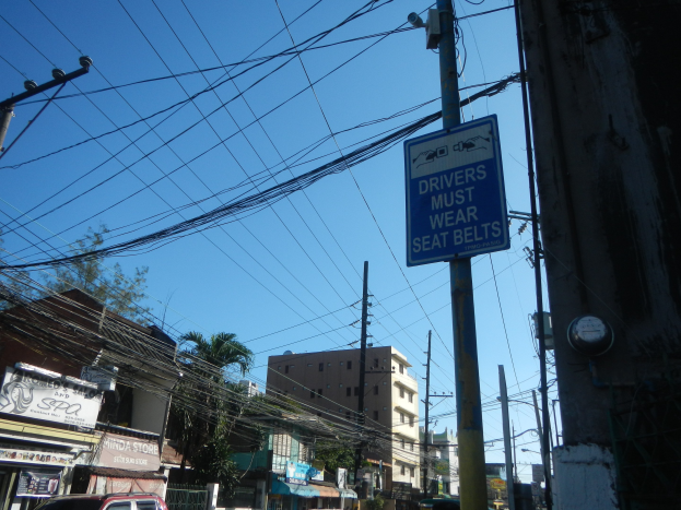 Stadtstraße mit fahrenden Autos, Strommäste mit Drähten, Gebäude mit Fenstern, Bäume, Namensschilder und ein "Fahrer Müssen Sitzgürtel Tragen"-Schild an einem Strommast vor einem sichtbaren Himmel.