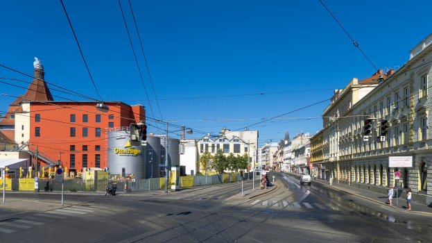 Eine Straßenszene in einer Stadt mit Gebäuden, Straßenlaternen, Verkehrszeichen, Fahrzeugen, Fußgängern, Bäumen und einem Himmel als Hintergrund.