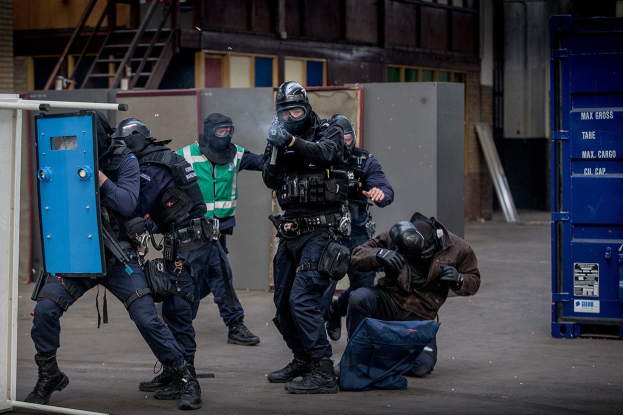 Eine Gruppe von Polizisten in Einsatzausrüstung steht vor einem Gebäude in Formation, einige halten Waffen, mit einer Person im Vordergrund und einem blauen Objekt mit Text auf der rechten Seite.