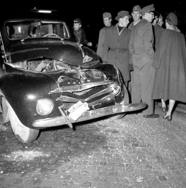 Ein Schwarz-Weiß-Bild eines verunglückten Autos am Straßenrand mit eingedellter Haube und zersplitterten Fenstern, umgeben von einer Gruppe besorgter Menschen.