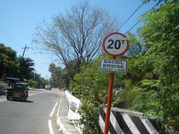 Eine Straße mit Fahrzeugen, ein Geschwindigkeitslimit-Schild, eine Trennlinie, Bäume, Strommasten mit Drähten und einen klaren blauen Himmel.