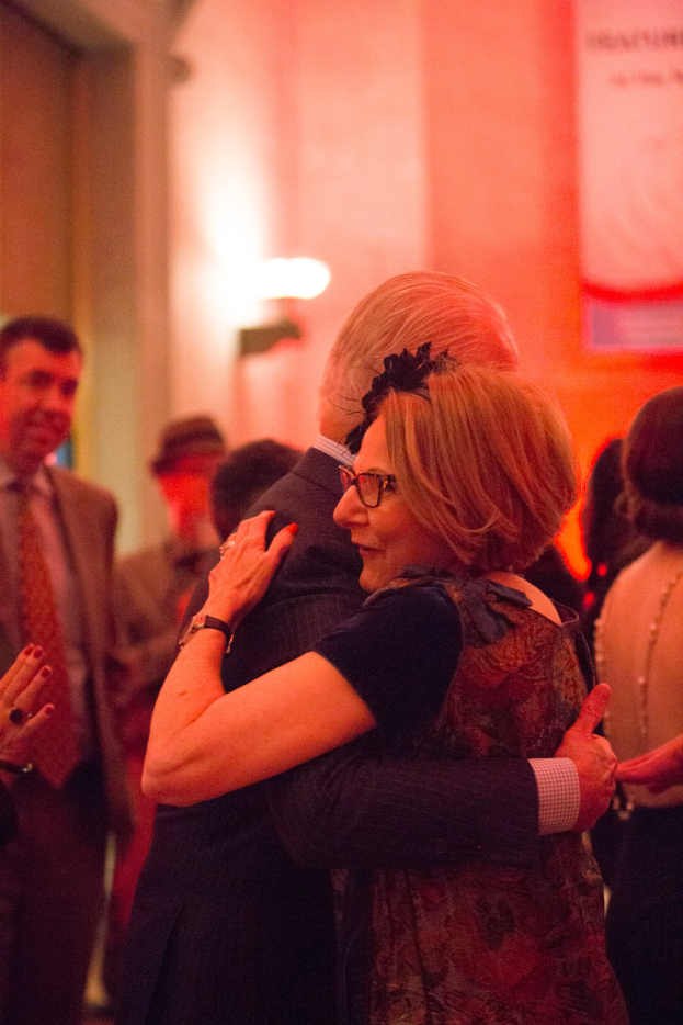 Zwei Menschen umarmen sich herzlich auf einer Party, umgeben von anderen im Hintergrund mit einer beleuchteten Wand.