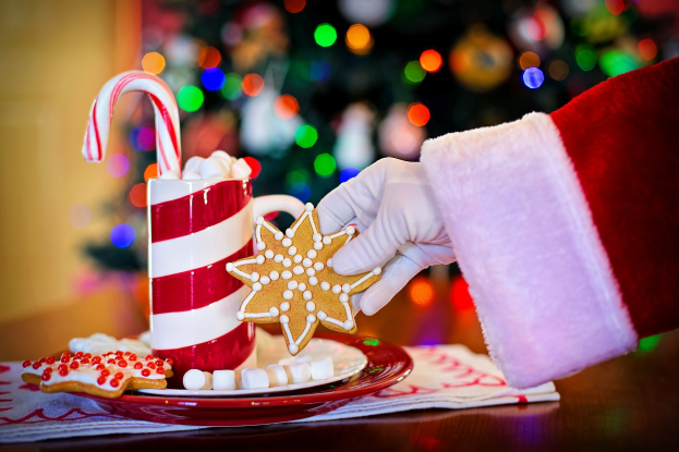 Eine Person mit einem Santa Claus Hut hält einen Becher heißer Schokolade und einen Bonbonstängel in der Hand und hat einen Teller mit Plätzchen und ein Tuch auf dem Tisch vor sich. Im Hintergrund sieht man einen Weihnachtsbaum und eine Wand.