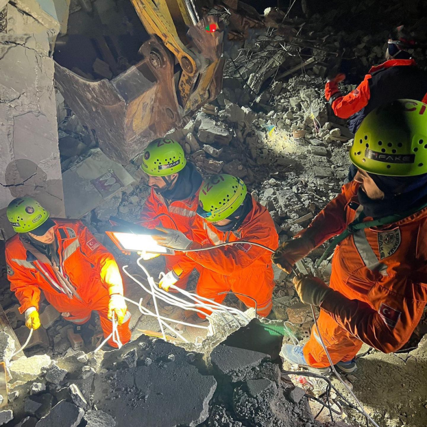 Eine Gruppe von Menschen in orangen Uniformen und grünen Helmen bei der Arbeit an Schutt mit einem Kran im Hintergrund, die Werkzeuge und Materialien halten.