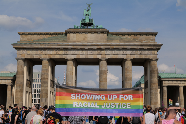 Eine Gruppe von Menschen hält eine "Racial Justice"-Schleife vor dem Brandenburger Tor in Berlin, wobei die Säulen und die Statue des Tors sowie Gebäude und ein bewölkter Himmel im Hintergrund zu sehen sind.