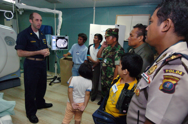 Eine Gruppe von Menschen in einem Krankenhauszimmer mit einem Mann, der in ein Mikrofon spricht, einem Bett mit einer Decke darauf links, medizinischen Geräten im Hintergrund, einem Röntgenmaschinenständer, einer Texttafel, einer Tür und Deckenleuchten.