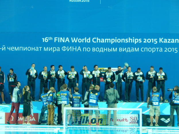 Gruppe von Menschen auf einem Schwimmbaddeck mit Kameras, einem Netz im Vordergrund und einem "FINA World Championships 2015 Kazan"-Banner im Hintergrund.