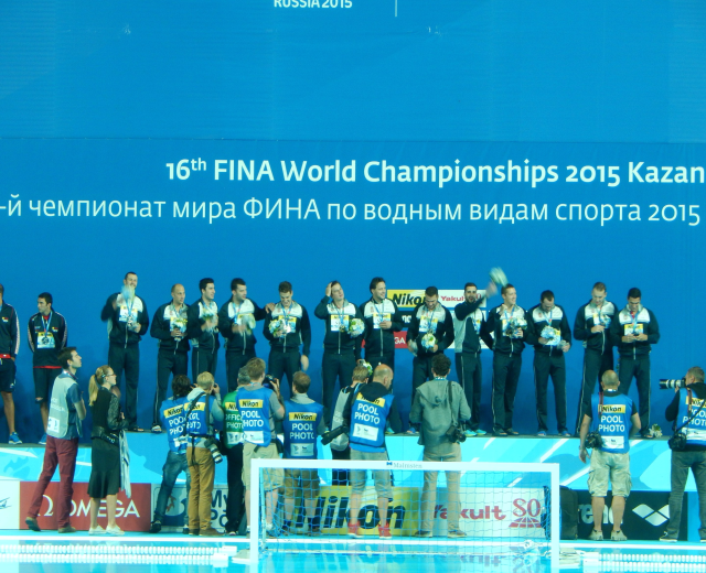Gruppe von Menschen auf einem Schwimmbaddeck mit Kameras, einem Netz im Vordergrund und einem "FINA World Championships 2015 Kazan"-Banner im Hintergrund.