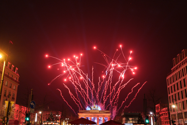 Eine nächtliche Straßenansicht am Silvesterabend in Berlin mit Gebäuden, Bäumen, Laternen, Verkehrszeichen, Schildern, Zelten, Menschen und einem Feuerwerk am Himmel.