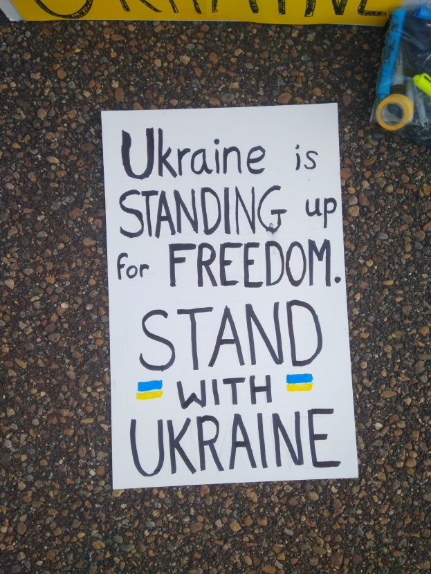 Weißer Papierbogen mit "Ukraine steht für Freiheit, steht mit Ukraine" darauf vor einem gelben Banner, mit einer Plastikhülle mit Gegenständen rechts.