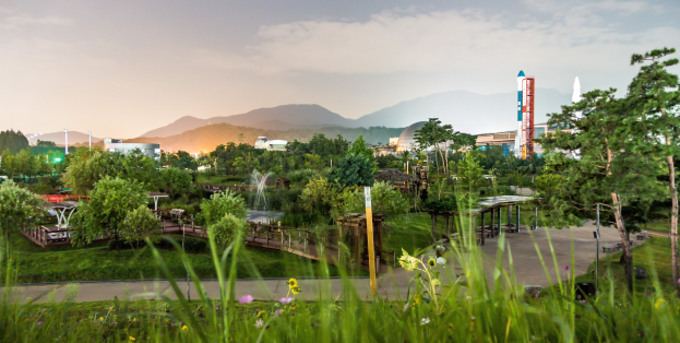 Ein Park mit grünem Gras, bunten Blumen, hohen Bäumen, einer Brücke, einem Springbrunnen, Laternenmasten und Gebäuden, mit Bergen und einem blauen Himmel im Hintergrund.
