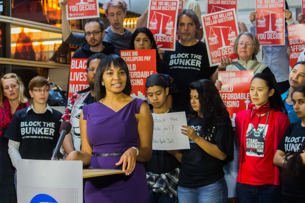 Frau mit Mikrofon an einem Rednerpult, die vor einer Menge mit Schildern spricht.