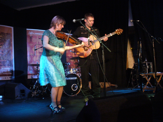 Ein Mann in schwarzem Hemd und Hose hält eine Gitarre in der Mitte der Bühne, mit einer Frau in einem blauen Kleid, die links von ihm eine Geige spielt, und verschiedenen Musikinstrumenten, einschließlich Lautsprechern und Trommeln, im Hintergrund.