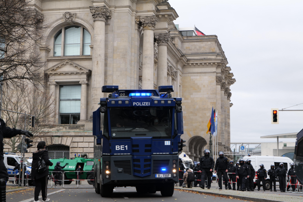 Eine Gruppe von Polizeibeamten steht vor einem großen Gebäude mit Fenstern, Säulen und Bögen, mit Fahrzeugen auf der Straße, einer Person mit einer Kamera auf der linken Seite und Bäumen, Verkehrsampeln, Flaggen und einem klaren blauen Himmel im Hintergrund.