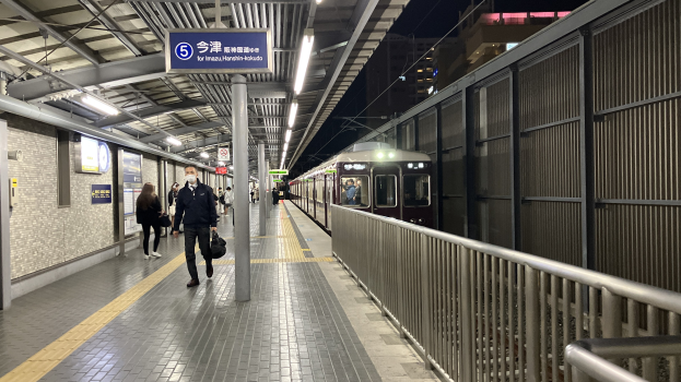 Ein Zug fährt auf einen Bahnsteig mit Menschen in der Nähe ein, ein Geländer auf der rechten Seite, Gebäude im Hintergrund, Bahnsteigbeleuchtung und ein Anzeigebrett.