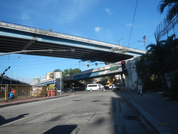 Eine Stadtstraße mit einer Fußgängerbrücke, Fahrzeugen, Fußgängern, einem Zaun, Topfpflanzen, Schildern, Laternen, Strommasten mit Drähten, Bäumen, Gebäuden und einer bewölkten Himmel.