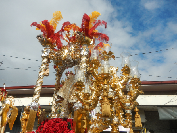 Ein großes goldenes und rotes Festwagen-Dekoration mit Blumen und anderen Schmuckstücken bei einem Faschingsumzug, mit einem Gebäude, Strommasten mit Drähten und einem bewölkten Himmel im Hintergrund.