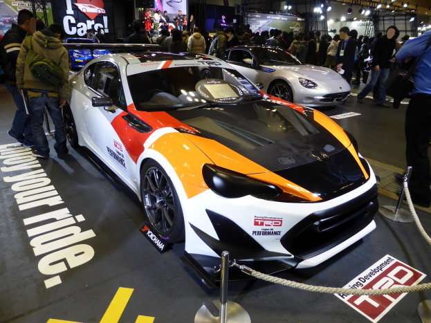 Eine Gruppe von Menschen steht um ein Rennfahrzeug herum, das auf der Tokyo Motor Show ausgestellt ist. Die Absperrung besteht aus Pfosten und Seilen, und es gibt Informationsschilder im Hintergrund unter beleuchteter Decke.