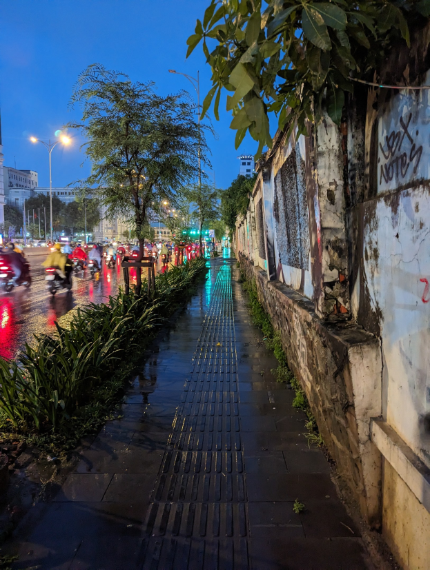 Stadtstraße bei Nacht mit Graffiti an der Seite, beleuchtet von Straßenlaternen, fahrende Kraftfahrzeuge, Bäume und Pflanzen am Straßenrand, Gebäude und ein sternenklarer Himmel im Hintergrund.