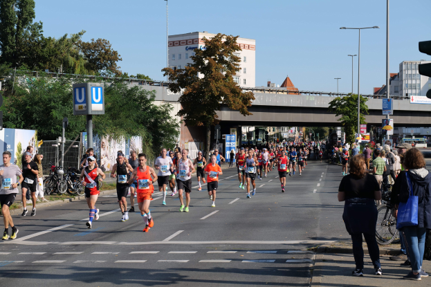Gruppe von Menschen, die bei einem Marathon auf einer baumgesäumten Straße laufen, mit Fahrrädern, Schildern, einem Zaun, einer Brücke, Gebäuden und einem klaren blauen Himmel.