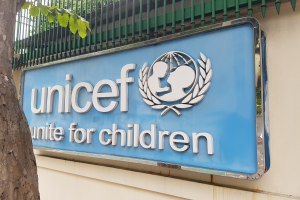 Das UNICEF-Logo ist auf der Wand eines Gebäudes zu sehen, mit einem Baumstamm links daneben und einer an der Wand befestigten Tafel, vor einem Hintergrund aus Bäumen und einem klaren blauen Himmel.