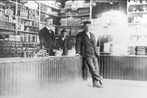 Drei Männer stehen an einem Tresen in einem Laden mit verschiedenen Gegenständen, einem buchgefüllten Gestell im Hintergrund, während eines Raubüberfalls.