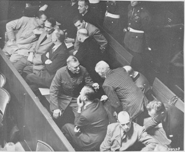 Ein Schwarz-Weiß-Bild einer Gruppe von Männern in einem Gerichtssaal, mit Adolf Hitler in der Mitte sitzend, umgeben von anderen Männern, einige stehend, und Stühle, die herumliegen.