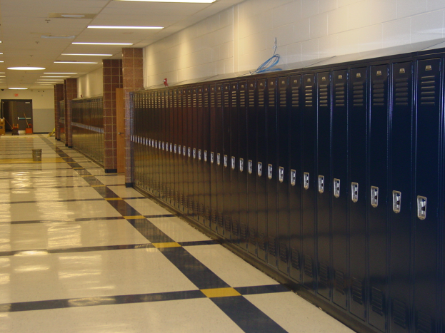 Eine lange Reihe von Spinden in einem Schulflur mit einem gefliesten Boden, einer Wand im Hintergrund, einer Decke mit Lampen oben und ein paar Gegenständen drumherum.