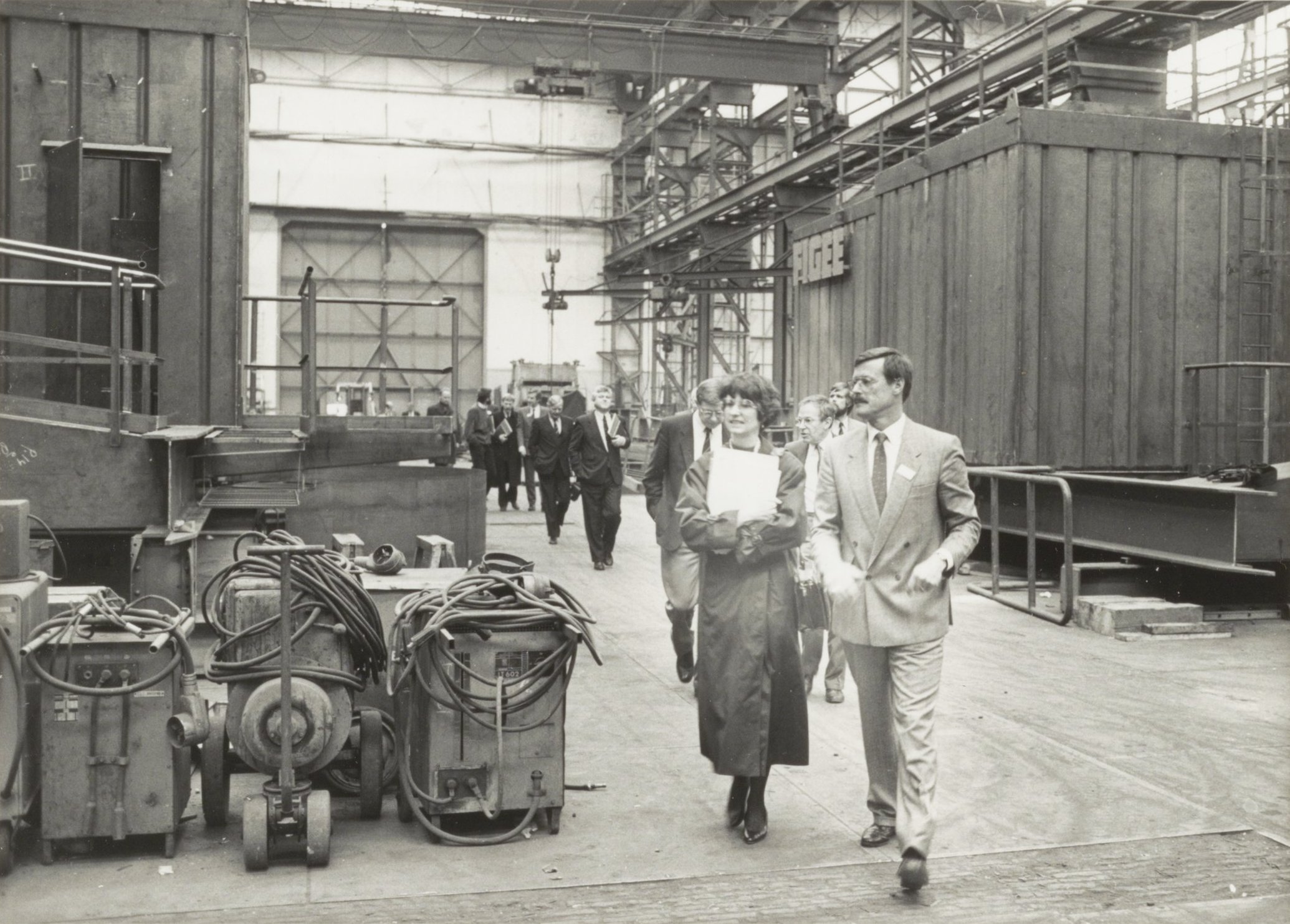 Eine Gruppe von Menschen in Anzügen und Schuhen geht durch eine Fabrik, mit Maschinen auf der linken Seite und einer Wand im Hintergrund.