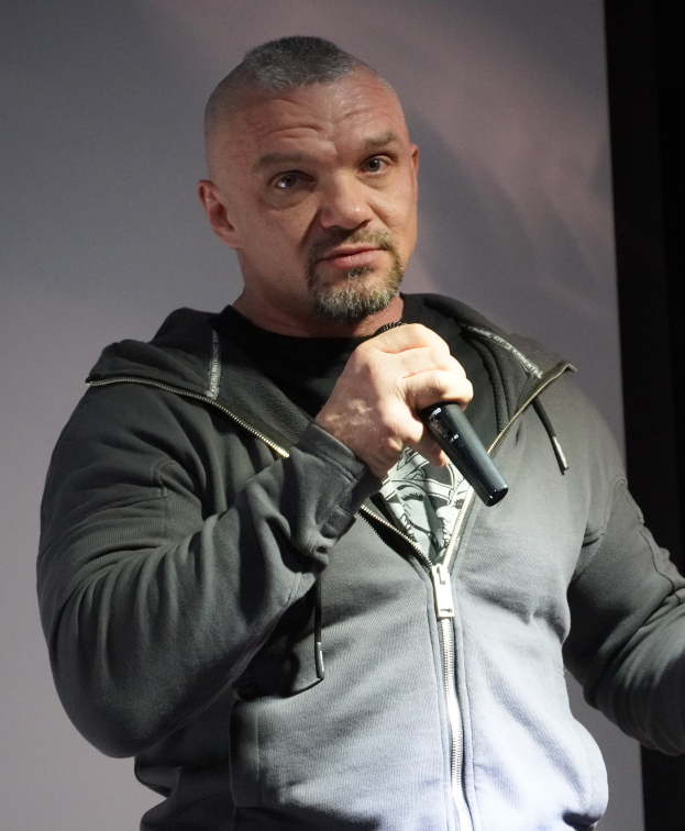 Ein Mann in einer schwarzen Kapuzenjacke und einem schwarzen Anzug mit einem weißen Hemd, zurückgekämmtes Haar und ein selbstbewusstes Lächeln hält ein Mikrofon, während er vor einer Wand steht.