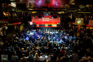 Eine große Menschenmenge sitzt auf Stühlen vor einer Bühne mit Bildschirmen und Bannern, beleuchtet von Scheinwerfern und umgeben von Geländern, voller Erwartung auf den Beginn eines Online-Marketing-Rockstars-Events, mit einem Wasserzeichen in der linken unteren Ecke.