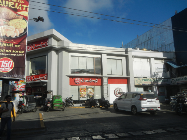 Ein weißes Gebäude mit einer Tafel, auf der 'Chowkint's Restaurant in Cebu City' steht, ist von Fahrzeugen auf der Straße, einem Fußgänger auf dem Gehweg, einer Ampel und Strommasten mit Drähten umgeben, unter einem bewölkten Himmel.