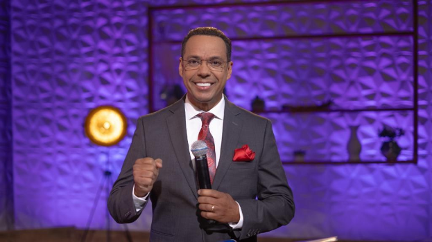 Stephen Colbert, Moderator von *The Daily Show with Trevor Noah*, steht auf einer Bühne und hält ein Mikrofon in der Hand, trägt einen Anzug, Krawatte und eine Brille, mit einer Lampe und einem Ständer im Hintergrund.