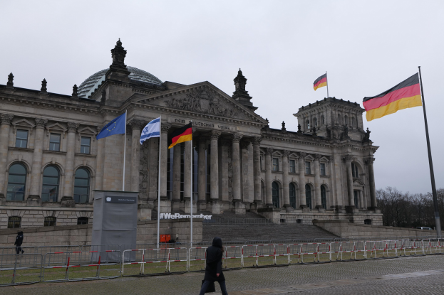 Eine Person, die vor dem Reichstaggebäude in Berlin, Deutschland, mit seinen Fenstern, Säulen, Bögen und Statuen, umgeben von Fahnenmasten und Geländern, unter Bäumen und einem klaren blauen Himmel geht.