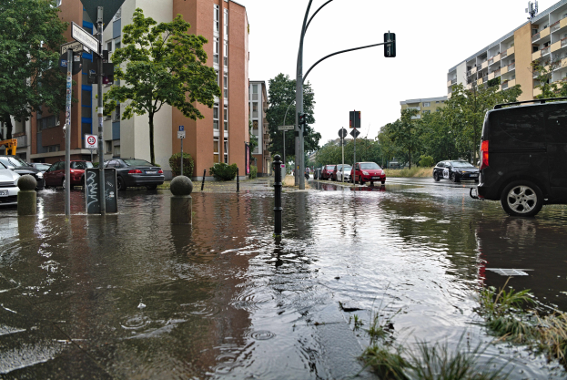 Eine überflutete Straße in Berlin, Deutschland mit parkenden Autos, Gras, Pfählen, Verkehrszeichen, Schildern, Bäumen und Gebäuden mit sichtbaren Fenstern im Hintergrund.